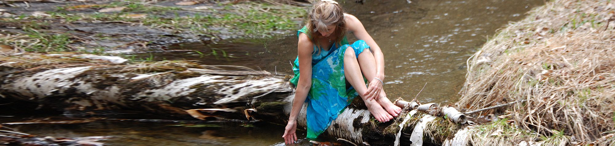 Debbie sitting on a log reaching out to the water in a stream