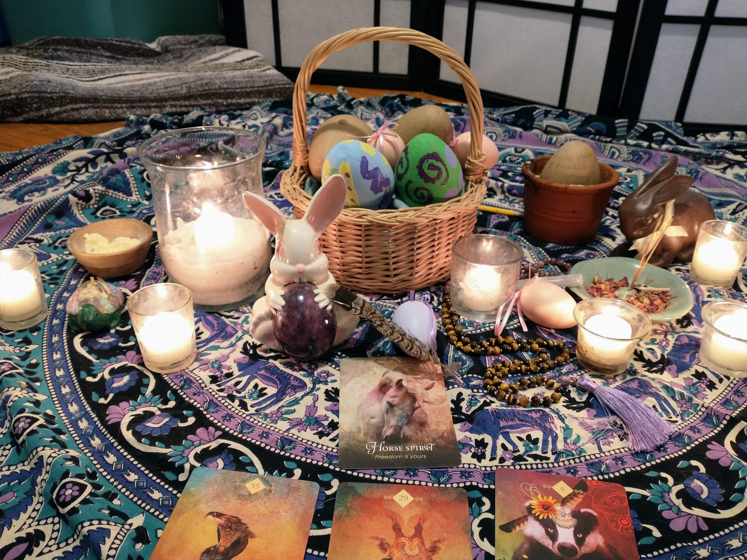 An altar laid out on a colored tablecloth with candles, ceramic bunnies, and a basket of colored eggs